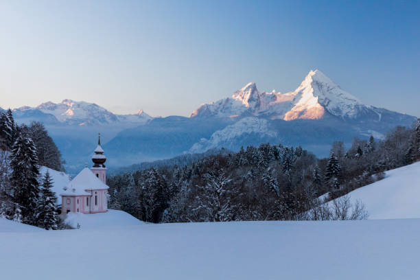Bird's-eye photo of a white church with a blue onion dome, red roofs, and snow-dusted buildings in a small European village. The image captures streets, a curved road, and a serene winter scene.