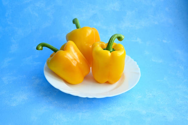 A vibrant close-up of three fresh, juicy yellow bell peppers with their stems facing up, placed on a white porcelain plate against a blue background. This bright and colorful image emphasizes the natural beauty and freshness of the vegetables, making it perfect for culinary, food photography, and healthy lifestyle themes.