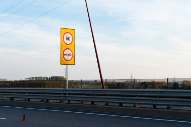A road sign on a highway bridge indicating a 60 km/h speed limit and a no overtaking for cars. The sign is mounted on a yellow background, standing along a modern bridge with red railings and lampposts, against a partially clear sky.
