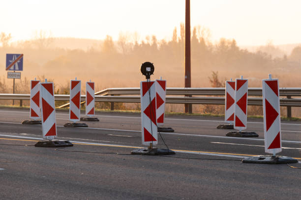 A row of red and white road construction barriers set up on a highway, illuminated by the gentle light of sunrise. The scene is enhanced by the warm morning atmosphere and misty background, creating a tranquil setting.