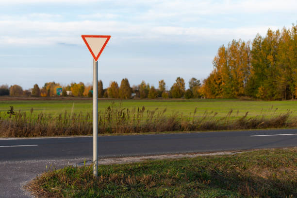 A triangular yield sign positioned along a rural road, set against an open field and autumn trees. The sign is clearly visible against the serene countryside, providing guidance for approaching drivers.