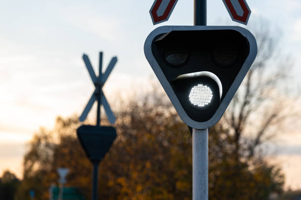 A close-up of an illuminated railroad crossing signal, showing a bright warning light with a crossbuck sign in the background. The scene is set at dusk, with soft lighting and a hint of autumn foliage in the background.