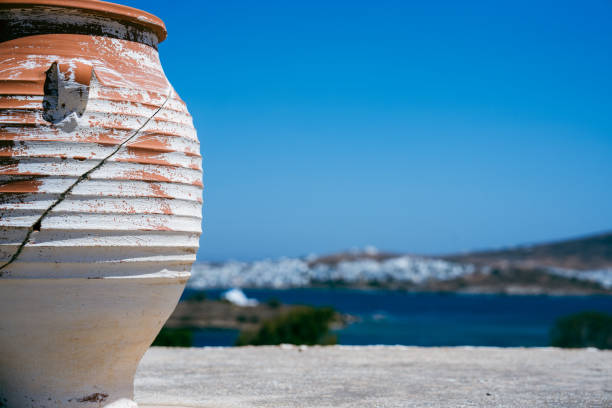 White and orange urn sits on a concrete wall. In the background behind the pot is an ocean and the white buildings of a small Greek island town.