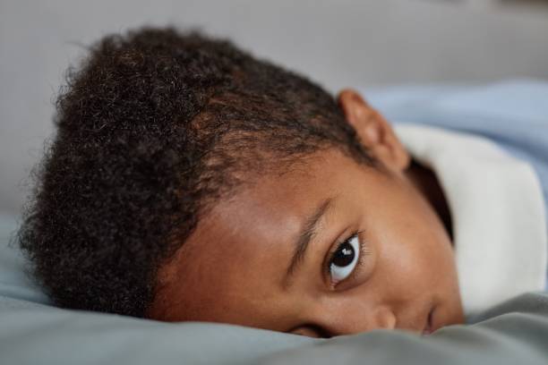 istock Tired Little Black Boy Looking at Camera while Resting on Bed at Home 2184789142
