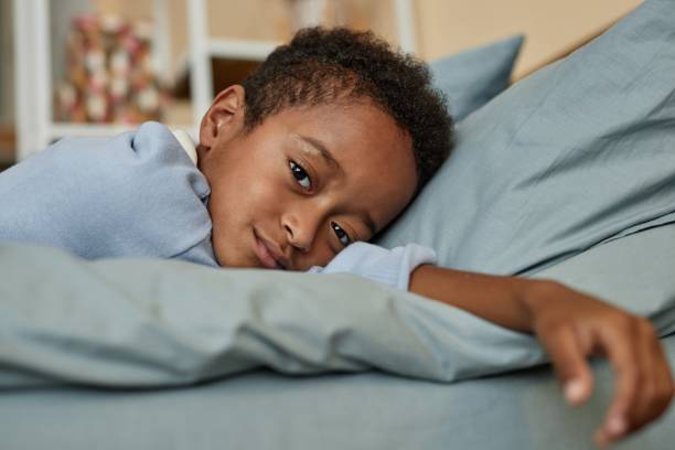 istock Tired Black Boy Resting on Pillow while Looking at Camera in Bedroom 2184789219