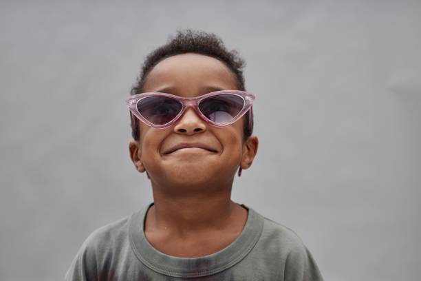 Studio shot of stylish little Black boy wearing sunglasses smiling with joyful expression on face while posing in studio against blurred gray background, copy space