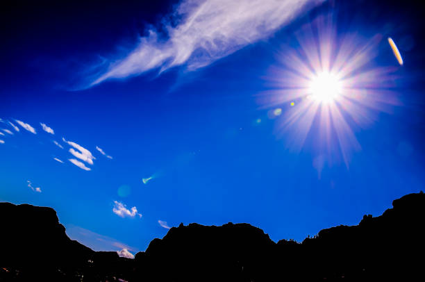 A bright sun is shining in the sky, casting a warm glow over the mountains. The clear blue sky is dotted with fluffy white clouds, creating a serene and peaceful atmosphere