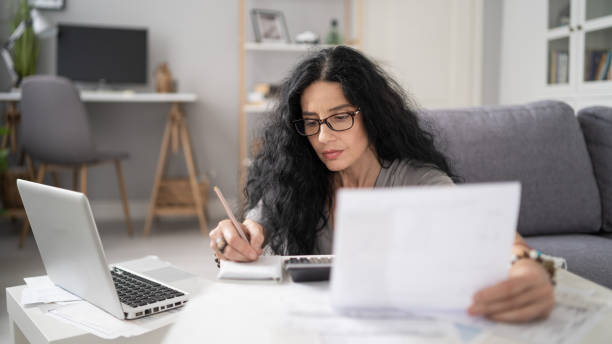 istock Concentrated woman in glasses writing notes, holding papers, cozy home office setup. 2184810336