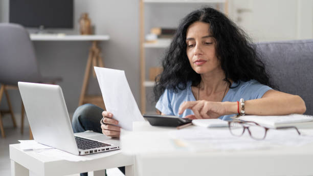 istock Focused woman checking calculations with documents and tech gadgets at her modern home. 2184810343