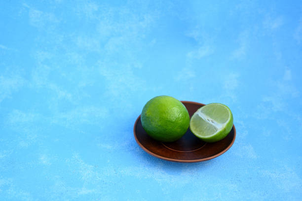 Two fresh limes on a small brown clay plate set against a vibrant blue background, with one lime cut in half to reveal the juicy, bright green interior. This arrangement highlights the natural, rustic appeal of citrus fruit, ideal for food presentations, healthy eating visuals, and tropical-themed projects.