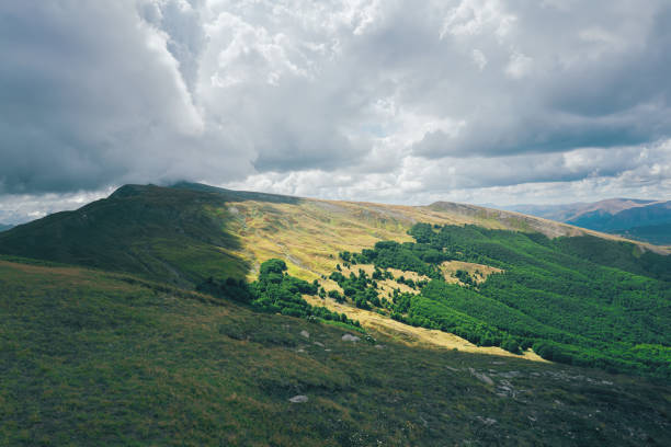 A scenic view of a valley on Macera della Morte, part of the Monti della Laga in Ascoli Piceno. The landscape is dotted with patches of sunlight breaking through heavy clouds, illuminating the green areas. The wide-angle shot enhances the vastness of the landscape, contrasting shadowed hills with sunlit slopes.