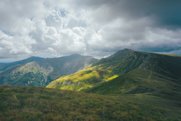 Vibrant green hills unfold into the distance, showcasing intricate terrain patterns and valleys. Overhead, clouds create soft, diffused lighting that enhances the texture and color of the landscape. A wide dynamic range highlights the variations in shadow and light, creating a balanced and immersive image.
