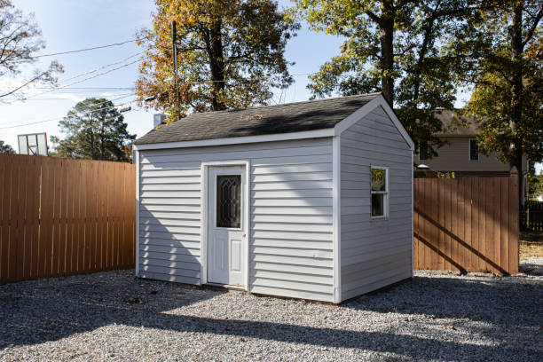 A quaint wooden shed with white siding stands on gravel, enclosed in a fenced yard, surrounded by mature trees, providing storage and a rustic charm to the landscape.