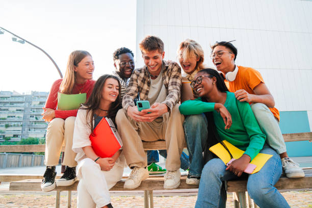 istock Group of real young people having fun together with a cellphone, siting on a bench at university campus. Multiracial friends watching the social media on a smartphone. Teenage students using a phone 2184850163