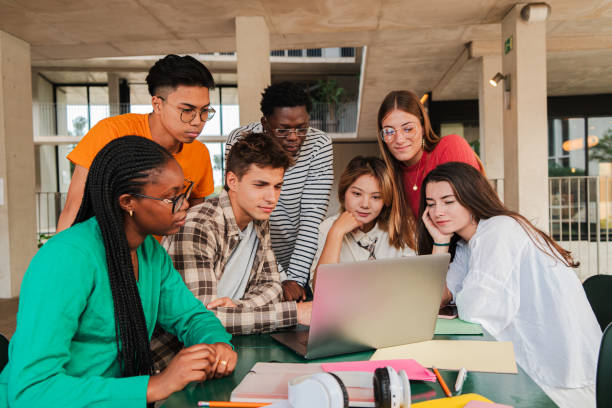 istock Large team of international young students using a laptop, collaborating on college research task in university campus library. Cooperation of teenagers finding data on notebook for a high school task 2184850276