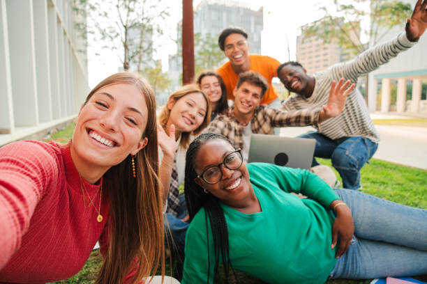 istock Blonde female student smiling taking a selfie with her classmates sitting at university campus lawn. Big group of teenagers laughing and looking at camera and posing for a portrait at the high school 2184854308