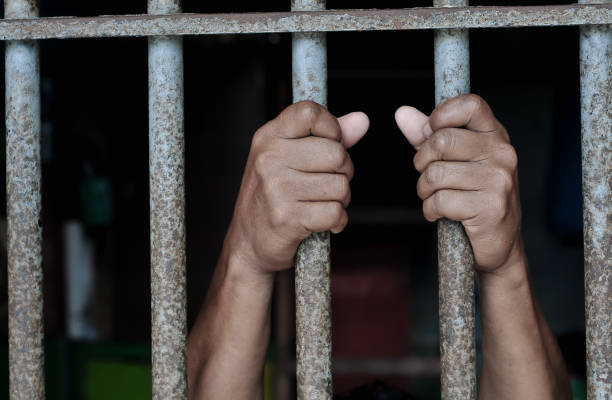 istock Hands of a prisoner behind prison bars on black background 2184889923