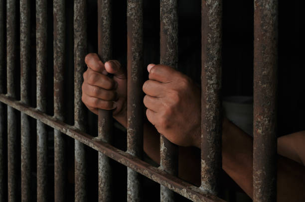 istock Hands of a prisoner behind prison bars on black background 2184889943