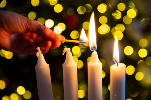 Hand lighting second candle on Advent wreath with four pillar candles. Christmas tree with twinkling lights on background. High quality photo