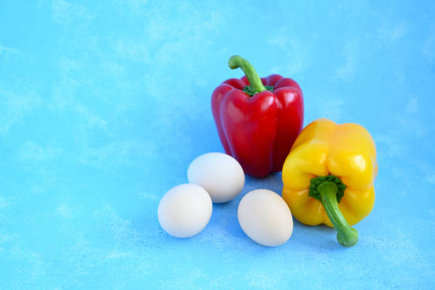 A vibrant still-life composition featuring a red bell pepper, a yellow bell pepper, and a rustic brown clay plate holding three white eggs. The setup is placed against a bright blue textured background, creating a striking color contrast. This minimalist and colorful arrangement highlights the freshness and simplicity of natural ingredients, making it ideal for themes related to healthy eating, organic food, culinary art, and farm-to-table concepts.