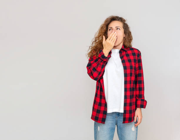 istock Curly woman in red shirt yawns against plain grey background in studio. 2184912789