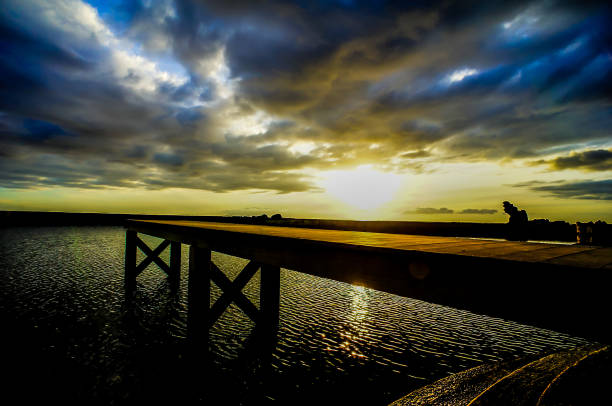 A pier is shown with a beautiful sunset in the background. The sky is cloudy and the sun is setting, creating a serene and peaceful atmosphere