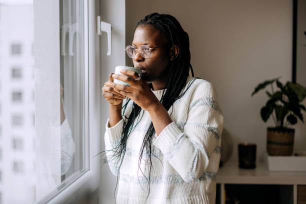 istock Casually clothed woman drinking coffee while standing next to window at home 2184923693