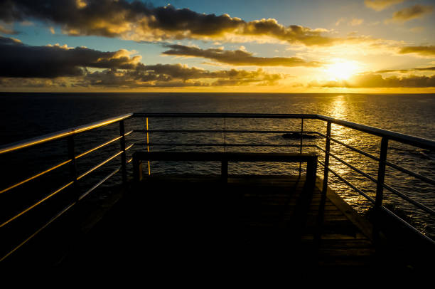 A pier overlooking the ocean with a sunset in the background. The sky is filled with clouds and the sun is setting
