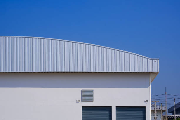 istock Factory warehouse building with automatic shutter doors on white concrete wall and aluminum dome roof against blue sky background 2184955735