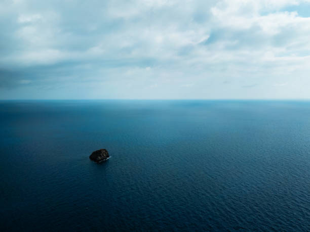 A vast expanse of calm ocean with a lone rock formation under a soft, cloudy sky. The scene evokes solitude, peace, and natural beauty. Ideal for themes of isolation and tranquility.