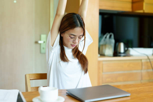 Asian woman with glasses raises arms to stretch while working at a wooden desk, wearing a casual white shirt, feeling bored and tired, taking a brief pause from her work in a quiet room. una mujer asiática con gafas levanta los brazos para estirarse mientras trabaja en un escritorio de madera, vistiendo una camisa blanca informal, sintiéndose aburrida y cansada, haciendo una breve pausa en su trabajo en una habitación tranquila.