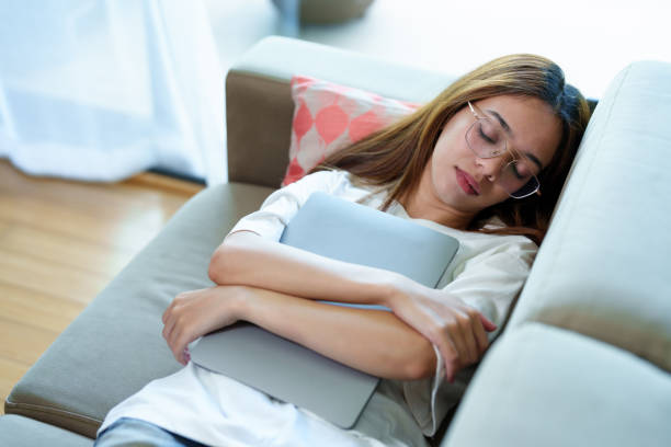 istock Asian woman with glasses asleep on a sofa while hugging a laptop, dressed casually in a white shirt, resting peacefully indoors after a tiring day of work, expressing exhaustion and boredom. 2184966801