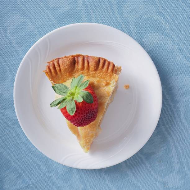 Slice of fresh apple pie with large red strawberry on top. A sweet treat viewed from directly above, on blue tablecloth background.
