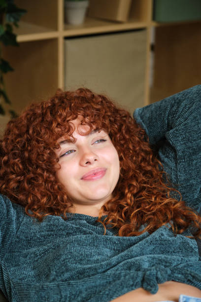 Serene young woman with curly red hair resting on a comfortable sofa with her hands behind her head, enjoying a moment of peace and relaxation