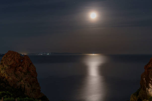 View of the full moon over the sea with the city in the background and rocks in the foreground.