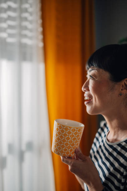 istock Asian woman holding a patterned mug looking out window in bright light 2185022271