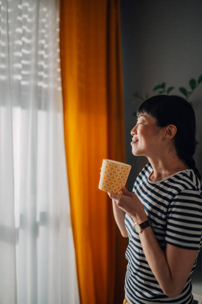 istock Asian woman holding a patterned mug looking out window in bright light 2185022279