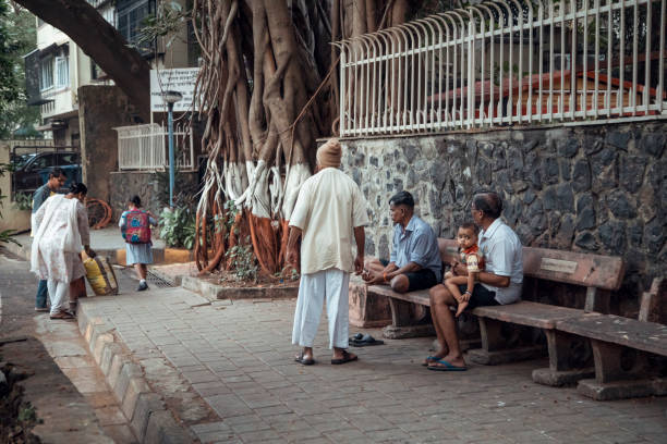 Mumbai, India - In a lively urban setting, a group of people engage in conversation while seated on benches. Children pass by, and a massive tree looms in the background, adding a natural touch to the environment.