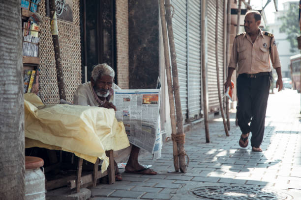 Mumbai, India - An elderly man sits on a chair, reading a newspaper under a yellow cloth. A passerby in a uniform walks by on a busy street lined with closed shops.