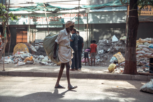 Mumbai, India - A man carries a sack walking along a crowded street. In the background, homeless children play near piles of trash. The scene captures the reality of poverty life midst rubbish and waste.