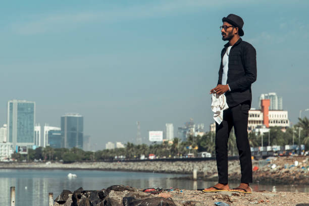 Mumbai, India - A man wearing a hat and casual attire stands on a rocky shoreline, gazing toward the city skyline under a clear blue sky. The urban backdrop contrasts with the tranquil water.