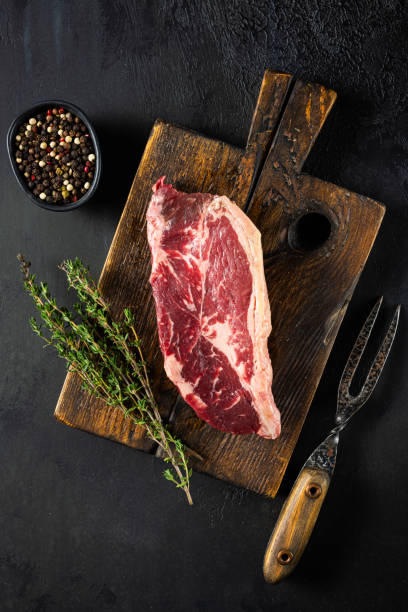 istock A raw ribeye steak placed on a rustic wooden board, accompanied by fresh thyme and a bowl of assorted peppercorns, ideal for culinary delights 2185075928