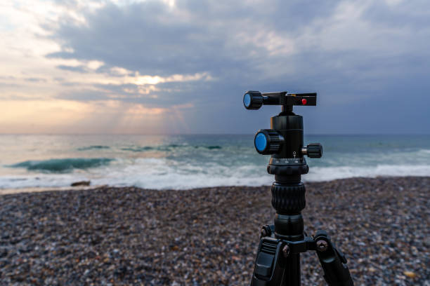 A calm beach scene at sunrise with a camera tripod on a pebble shore, capturing the peaceful waves and expansive sky. Ideal for concepts of photography, nature, and tranquility.