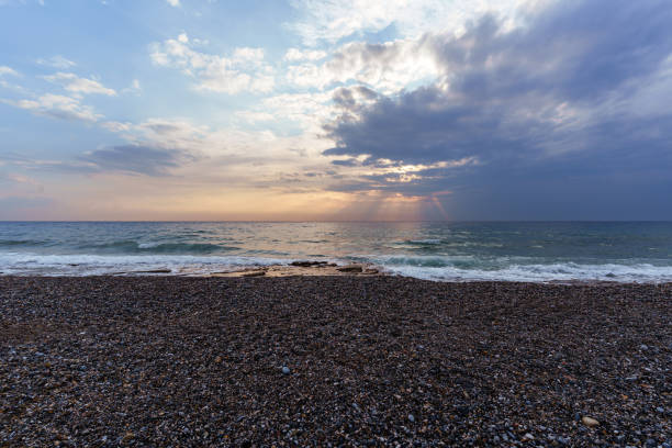 Tranquil scene of a pebble beach at sunset. The sky is filled with dramatic clouds and sun rays streak across the ocean, creating a peaceful and calming atmosphere.
