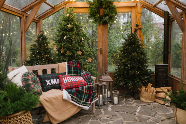 Cozy Christmas interior with a bench, plaid pillows, decorated Christmas trees, lanterns, and a warm festive atmosphere in a glass greenhouse.