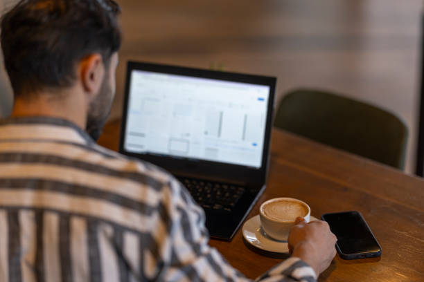 Young businessman works remotely in a cozy coffee shop, enjoying a cappuccino and staying connected on his smartphone, embodying the flexibility of remote work