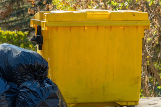 istock In the open air, a vibrant yellow garbage bin is positioned next to several black plastic bags, contrasting against the lush greenery of a well-kept landscape under clear blue skies 2185137990