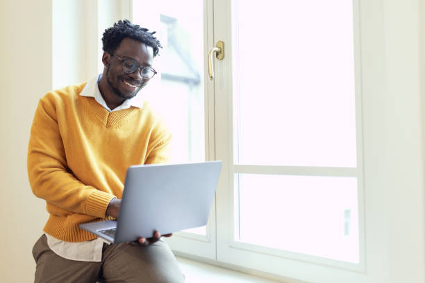 istock Smiling black business man using laptop while standing by window in office 2185142329