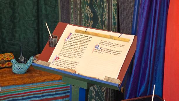 An open illuminated medieval manuscript rests on a wooden stand, surrounded by medieval-inspired decor, with colorful calligraphy and an ink pot with a quill, evoking a historical or artistic setting.