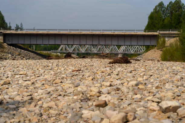 A view of two bridges, one for rail and one for vehicles, spanning a dry, rocky riverbed. The scene captures the industrial structures amidst nature, with no people present